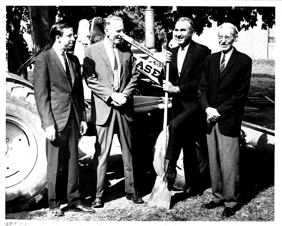 Four men in 1957 era clothing standing in front of construction equipment one of them is holding a shovel ground breaking ceremony